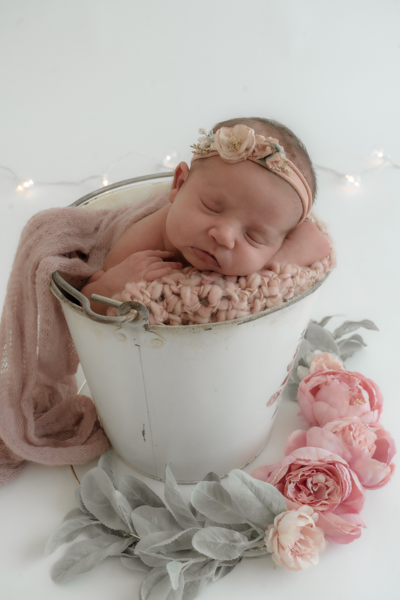 Newborn girl in a blush knit wrap resting in a white bucket with peonies