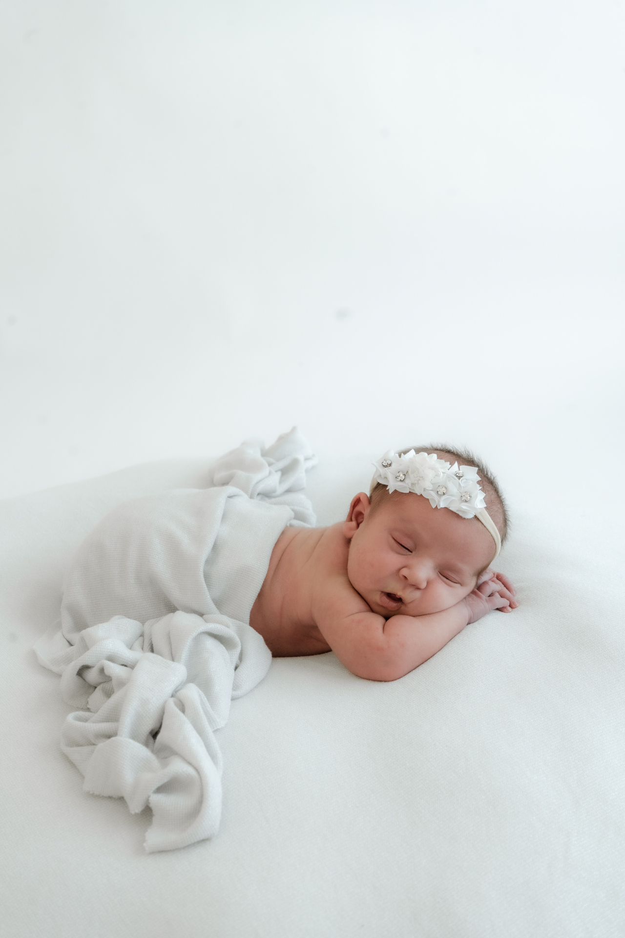 Newborn baby in a soft white wrap and floral headband on an ivory blanket