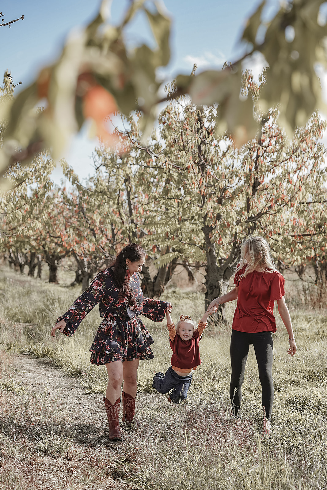 Mother and daughters swinging a toddler between them in a golden autumn orchard