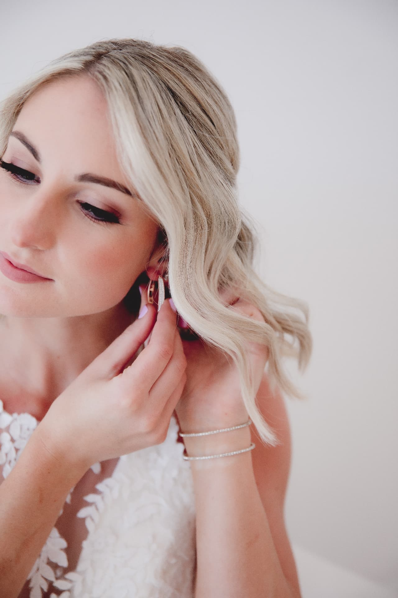 Bride with soft waved blonde hair fastening a diamond earring while wearing an ivory lace gown