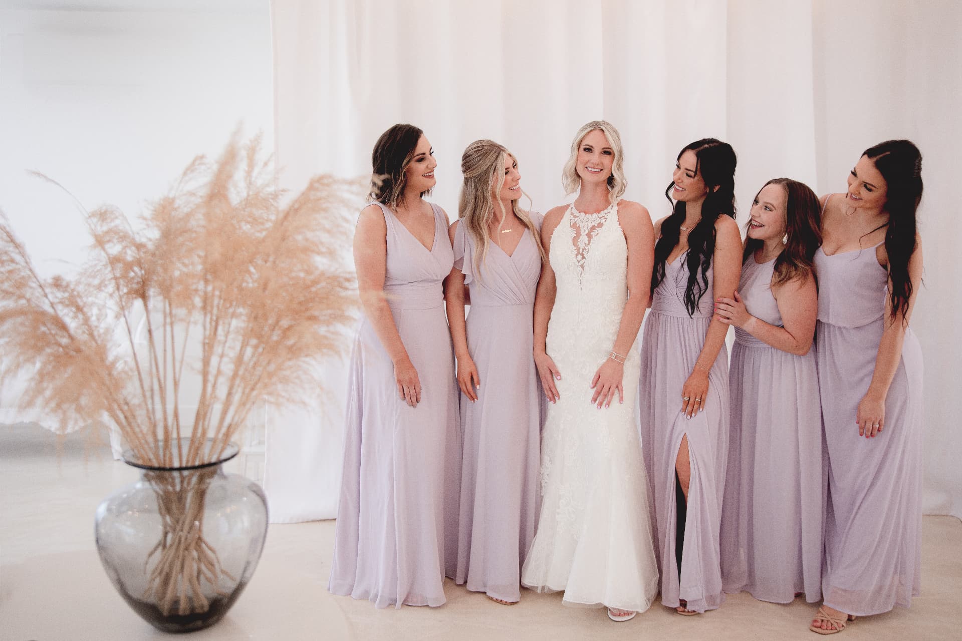 Bride in an ivory lace gown surrounded by her bridesmaids in lavender chiffon dresses inside the white-draped bridal suite at Glam Studios