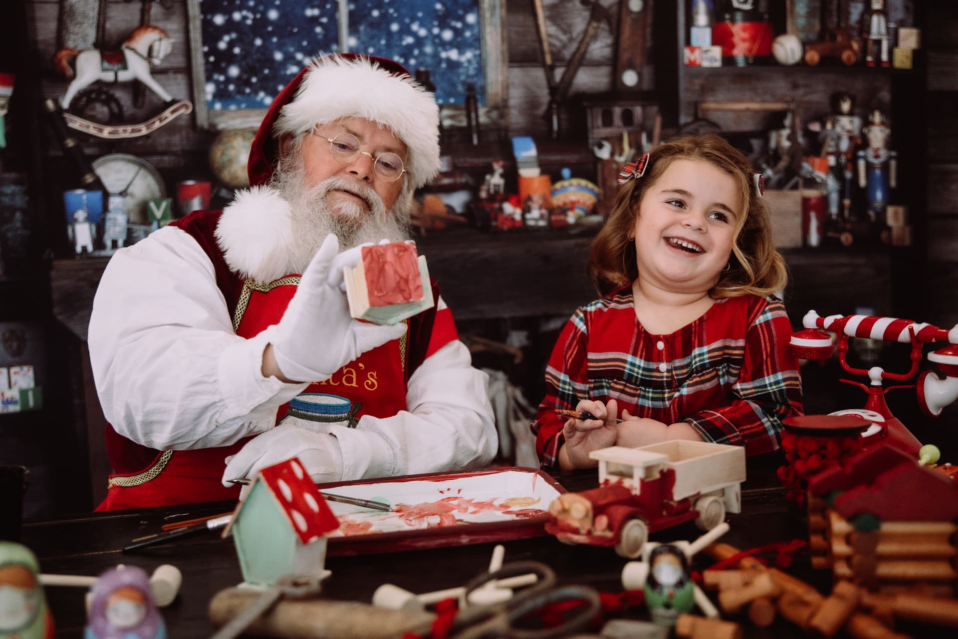Santa painting toys in his workshop beside a laughing little girl in red plaid