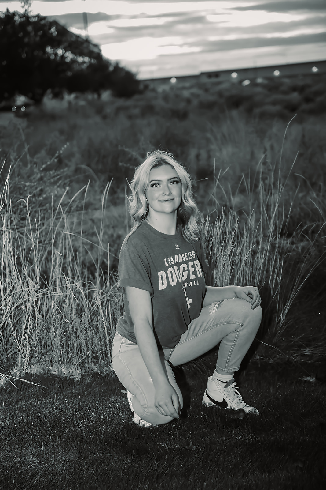 Black and white senior portrait kneeling in tall field grass at dusk