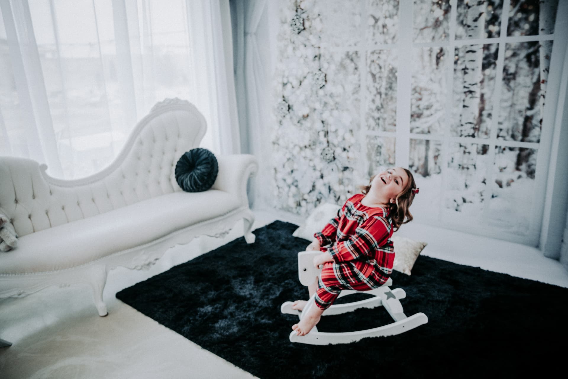 Girl in red plaid pajamas laughing on a white rocking horse in the winter white room